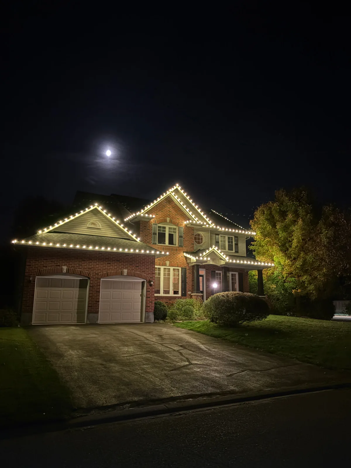 Brick home under a full moon with warm white holiday lighting outlining the rooflines